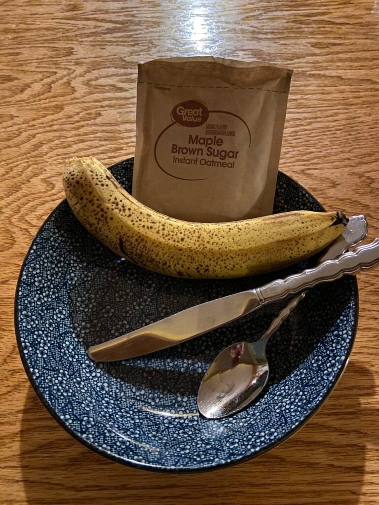 blue flower print pasta bowl sits on wooden table, dimly backlit. a brown envelope of instant oatmeal and a banana are at the back of the bowl. a spoon and knife are in the bowl, handles on the rim. 