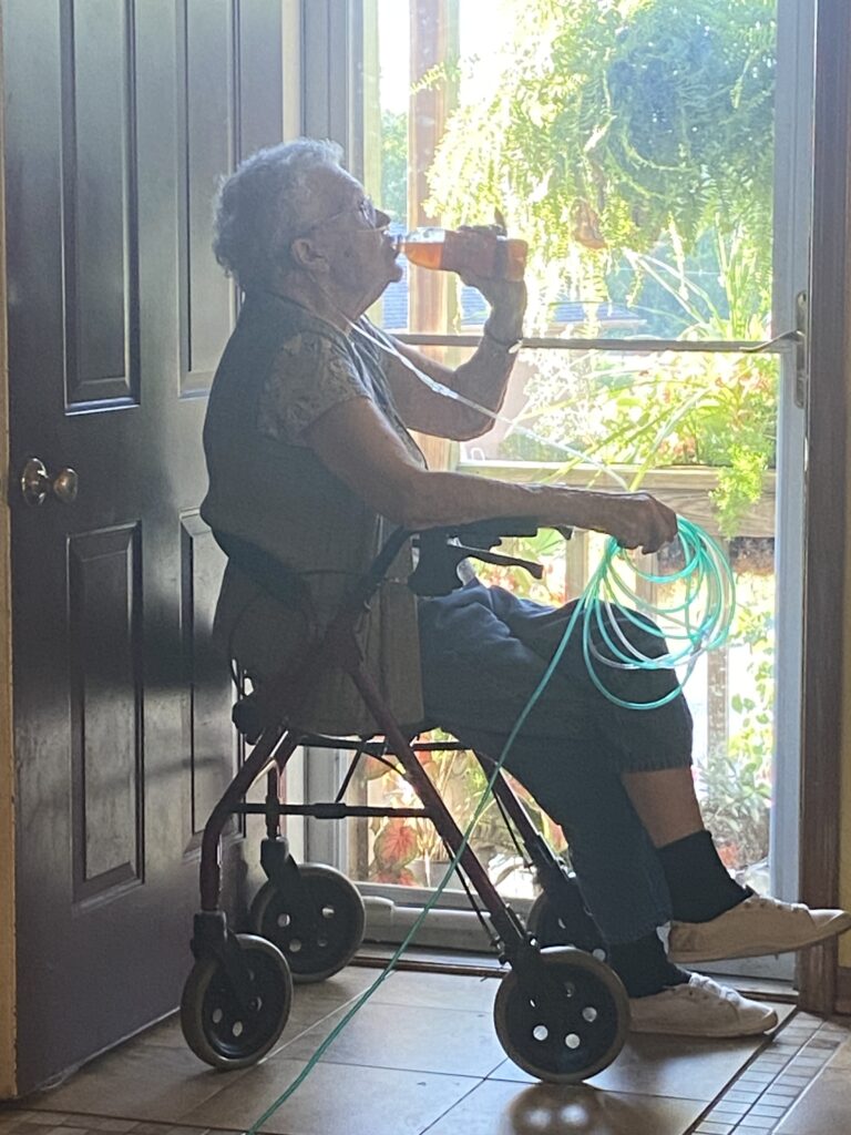 elderly lady sitting on walker in front of a glass front door. She has a bottle of Lipton tea in one hand, and oxygen tubing rolled up in the other. ferns and flowers line the porch, and you can see a wooden hand railing. Her shadow reflects off the front door behind her