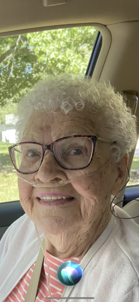 elderly lady sitting in car, kinky curls in her hair, glasses, and oxygen tubing in her nose. she is smiling and there are trees in the background
