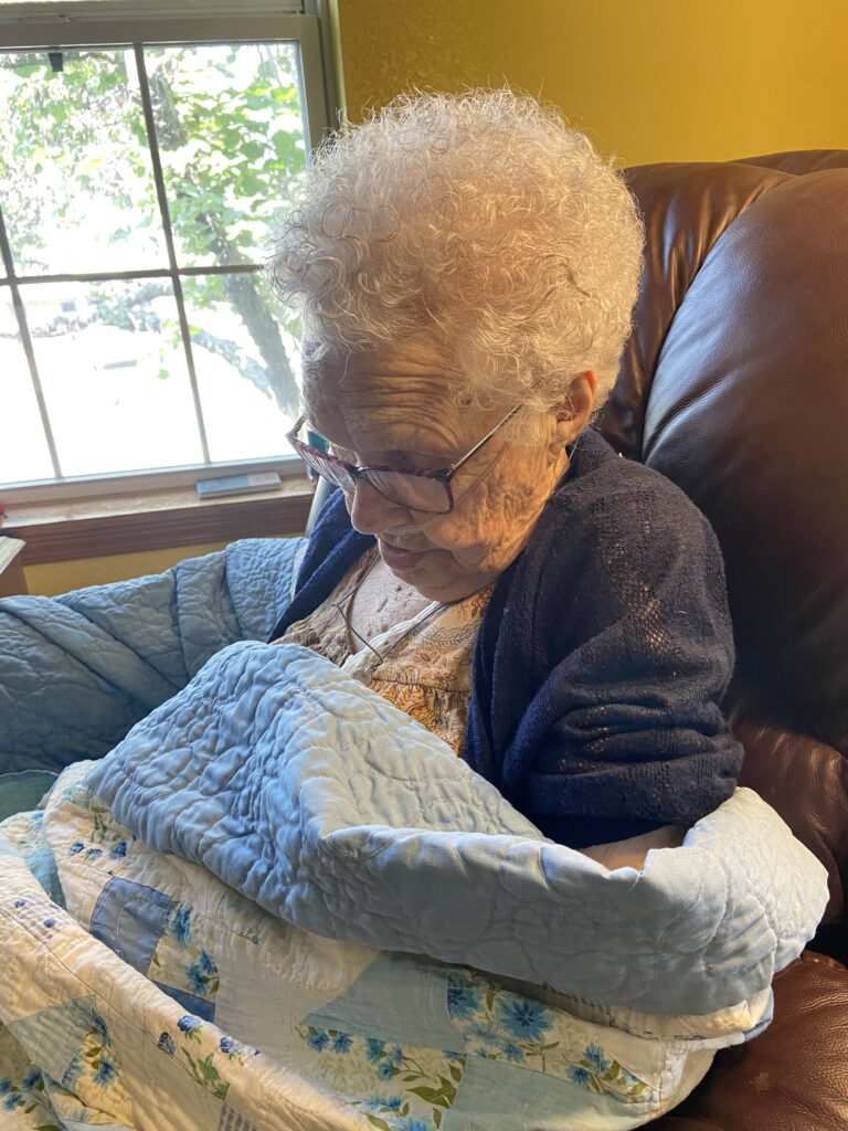 White haired lady sits in leather chair next to a window, wrapped in blue and white quilt, napping.