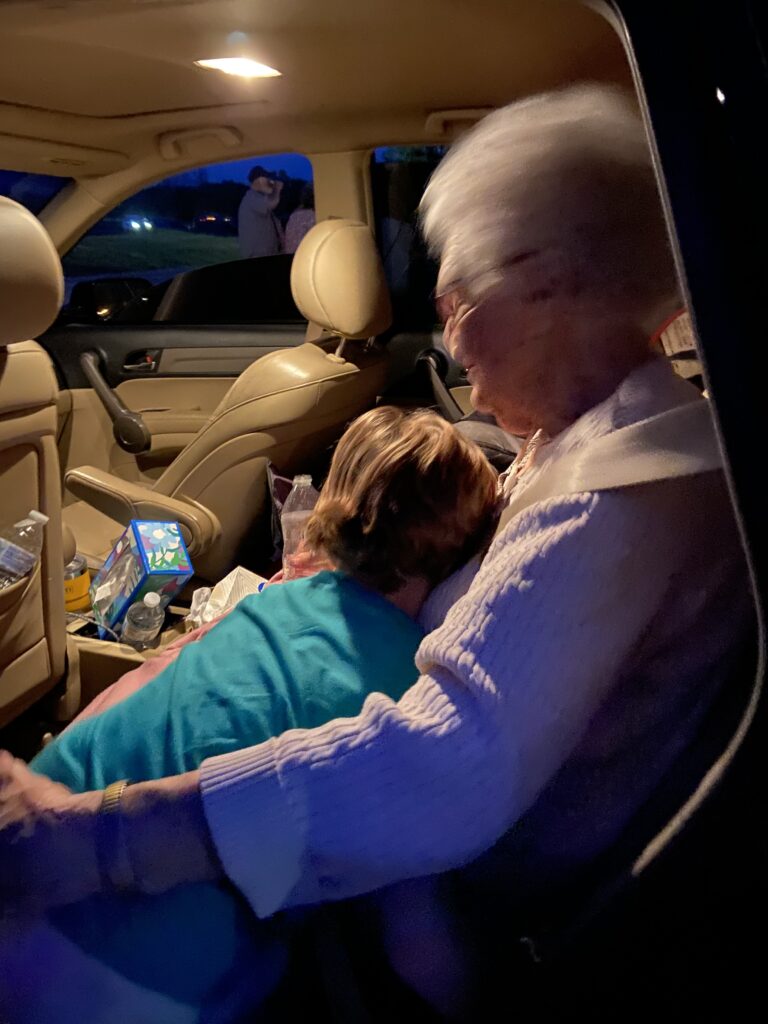 Old lady sitting in the back seat of a car, a child  in a turquoise shirt leans on her as if giving her a hug. a