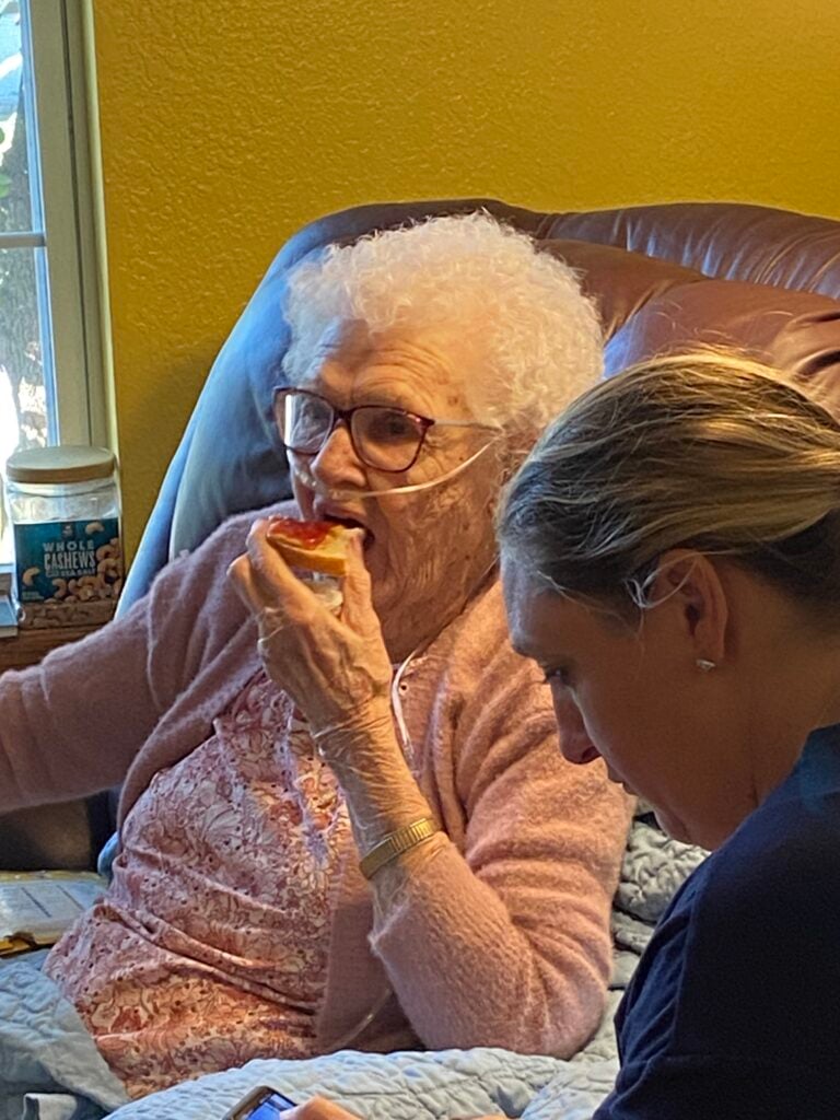 young lady with head bowed sits next to a white haired woman eating a piece of toast, sitting in a leather chair next to a window.