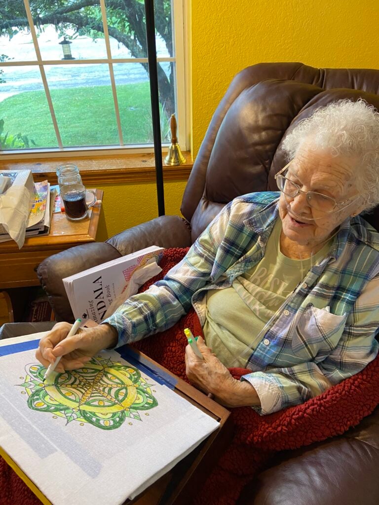 elderly woman sits in leather chair near a window, where spring green can be seen outside behind a a bird feeder hanging from a tree. a cup of coffee sits on a side table, and he lady is wrapped in a red blanket, coloring a green and yellow mandala on a large white background.