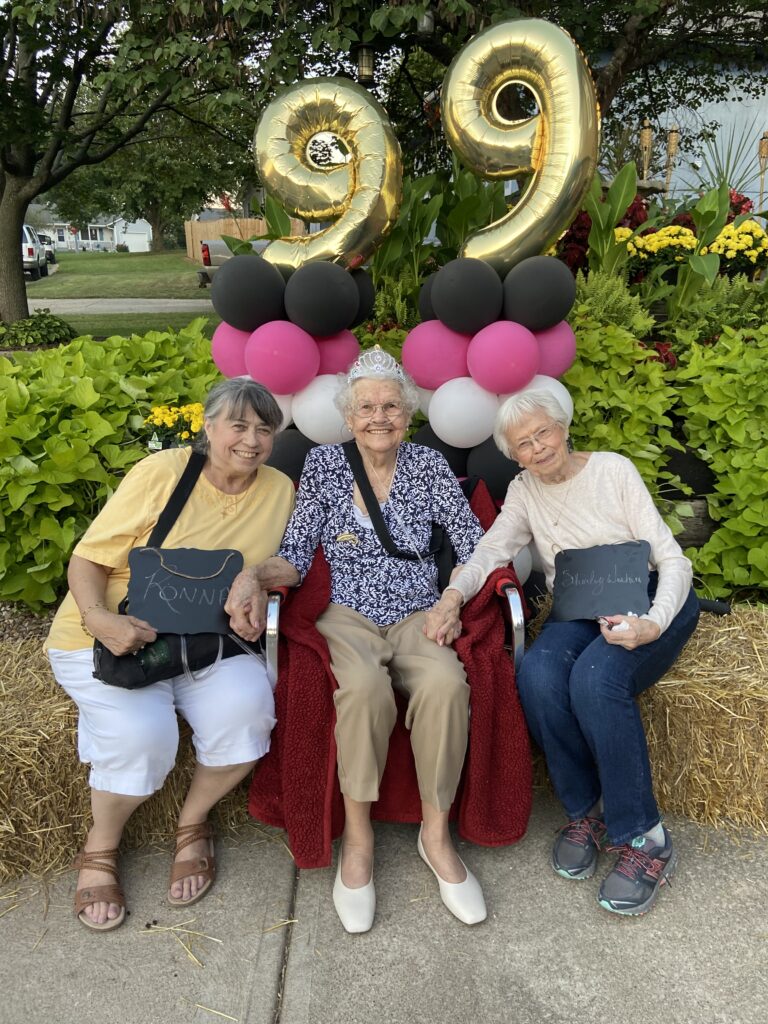 3 elderly ladies sit on hair bales, holding hands.the one on the right holds a small chalkboard saying Shirley; the one on the left says Ronna. They are smiling and the lighting appears to be dusk. giant gold foil 99 sits atop black, pink, and white balloon pillars, centered behind the lady in the center, who is wearing a silver princess tiara