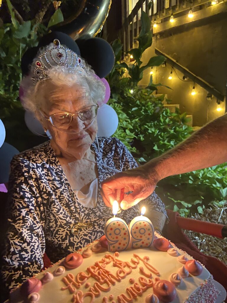 beautiful old lady sits holding a cake with numbers 99 as an arm and hand reach in from the right side to light the candle numbers with a cigarette lighter. The cake reads Happyy birthday in pink with white frosting and pink rosettes around the top edge and pink sprinkles on the side. The woman smiles fainly. she si wearing a princess tiara and has on glasses, and oxygen. behind her evening has turned to dusk and porch lights line a handrail . there are black pink and white blalloons just behind her head, and what appears to be the base of a giant gold foil #9. It is a happy picture