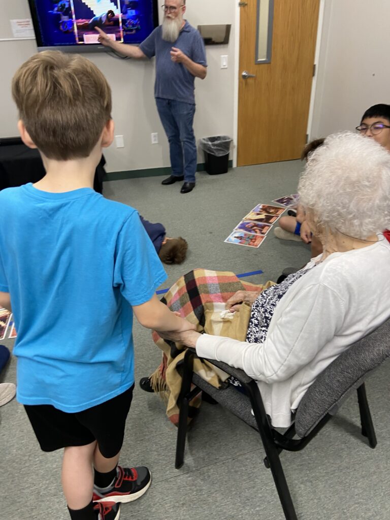 child in blue shirt holds the hand of an old lady who is sitting in a classroom chair. a man points to a tv as if teaching a class