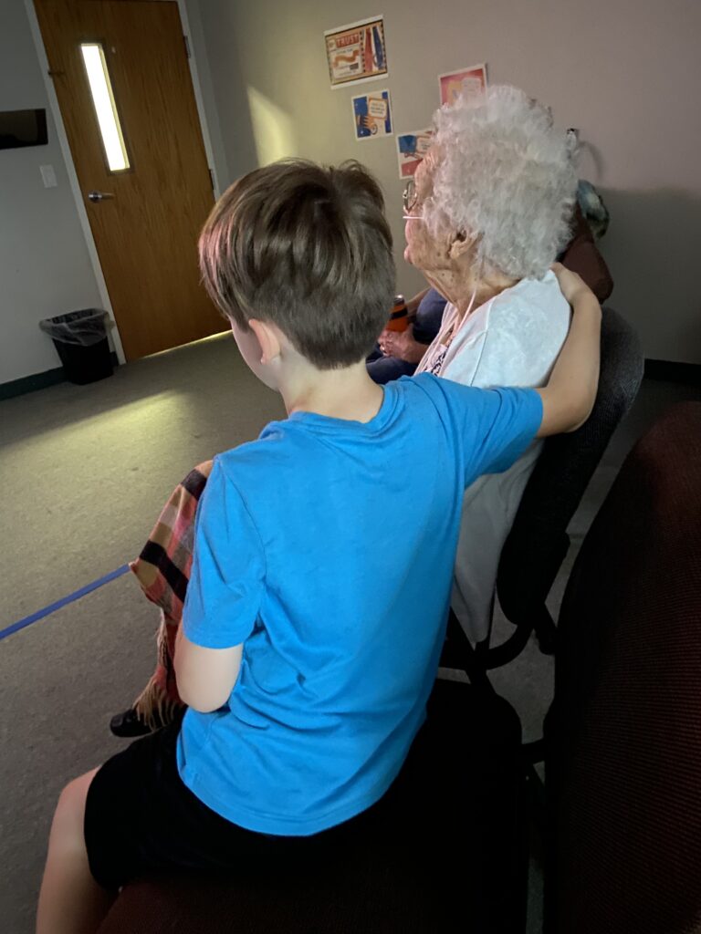 child in blue shirt sits next to old lady, his right arm around her shoulders
